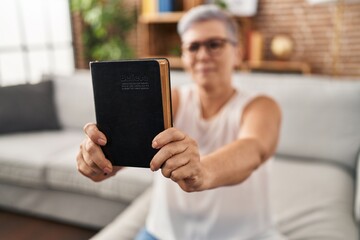 Middle age woman holding bible sitting on sofa at home