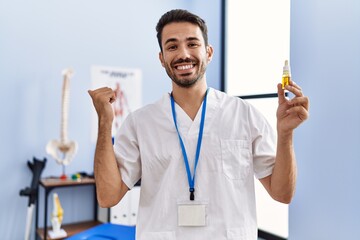 Young hispanic physiotherapist man holding cbd oil as pain relief pointing thumb up to the side...