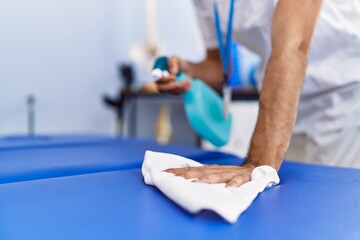 Young hispanic man wearing physiotherapist uniform disinfecting massage table at rehab clinic