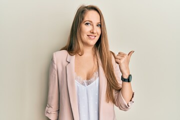 Young blonde woman wearing business jacket and glasses smiling with happy face looking and pointing to the side with thumb up.