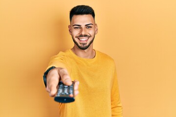 Young hispanic man with beard holding television remote control smiling with a happy and cool smile...