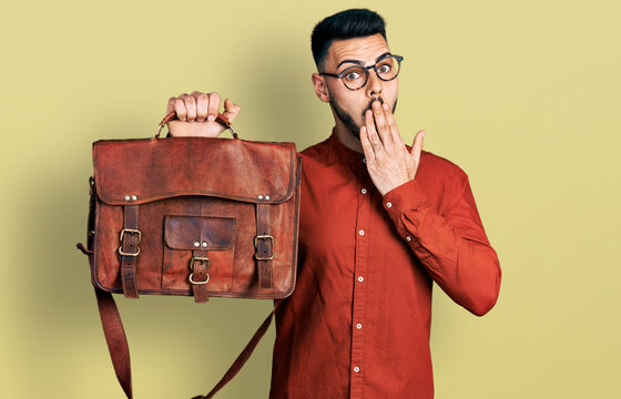 Young hispanic man with beard wearing leather bag covering mouth with hand, shocked and afraid for mistake. surprised expression