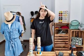 Young asian woman working as manager at retail boutique making fun of people with fingers on forehead doing loser gesture mocking and insulting. - obrazy, fototapety, plakaty