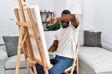 Young african man painting on canvas at home smiling cheerful playing peek a boo with hands showing face. surprised and exited