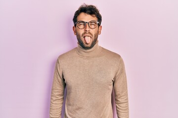 Handsome man with beard wearing turtleneck sweater and glasses sticking tongue out happy with funny expression. emotion concept.
