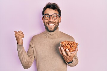Handsome man with beard holding peanuts pointing thumb up to the side smiling happy with open mouth
