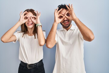 Young couple wearing casual clothes standing together doing ok gesture like binoculars sticking tongue out, eyes looking through fingers. crazy expression.