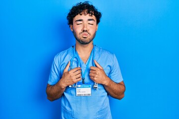 Young hispanic man wearing blue male nurse uniform puffing cheeks with funny face. mouth inflated...