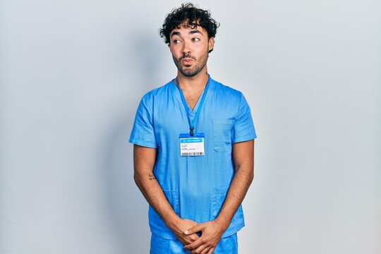 Young hispanic man wearing blue male nurse uniform smiling looking to the side and staring away thinking.