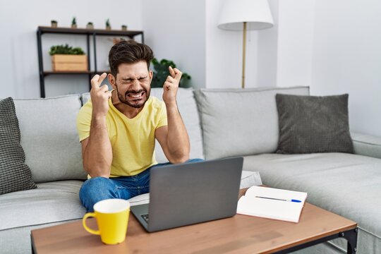 Young man with beard using laptop at home gesturing finger crossed smiling with hope and eyes closed. luck and superstitious concept.