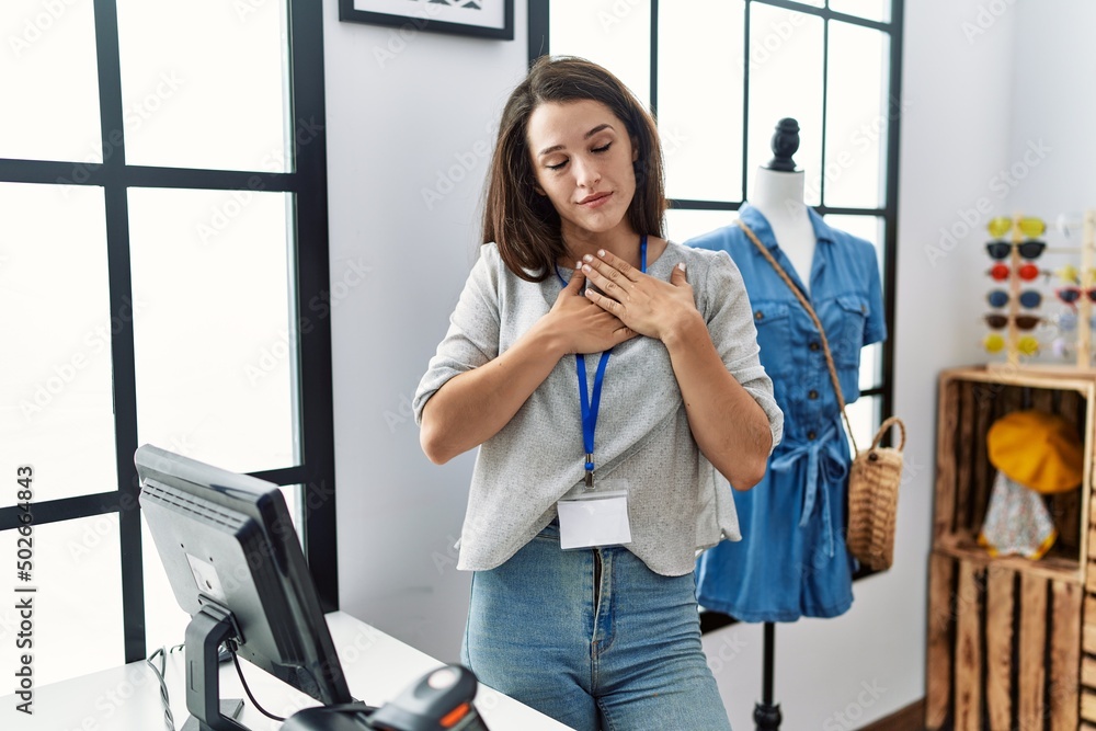 Poster young brunette woman working as manager at retail boutique smiling with hands on chest with closed e