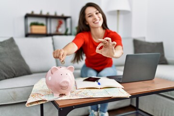 Obraz na płótnie Canvas Young brunette woman putting euro coin in piggy bank saving for travel doing ok sign with fingers, smiling friendly gesturing excellent symbol