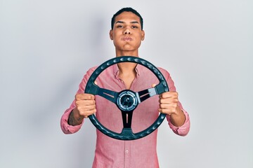 Young african american guy holding steering wheel puffing cheeks with funny face. mouth inflated...