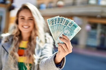 Young blonde woman smiling holding brazilian reals banknotes at the city