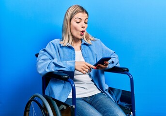 Beautiful caucasian woman sitting on wheelchair using smartphone scared and amazed with open mouth...