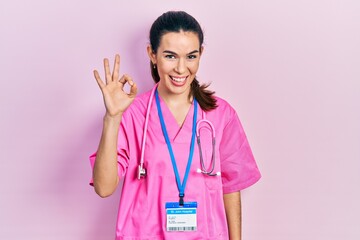 Young brunette woman wearing doctor uniform and stethoscope smiling positive doing ok sign with...