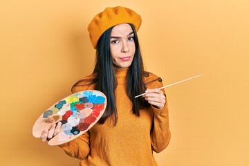 Young brunette woman holding paintbrush and palette wearing beret skeptic and nervous, frowning...
