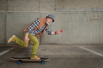 bearded senior man wearing round glasses and a bucket hat is riding a long skateboard in a grunge...