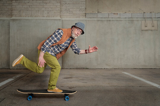 bearded senior man wearing round glasses and a bucket hat is riding a long skateboard in a grunge urban environment