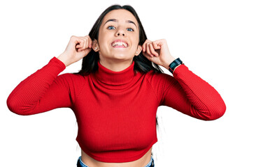 Young brunette teenager wearing red turtleneck sweater smiling pulling ears with fingers, funny...