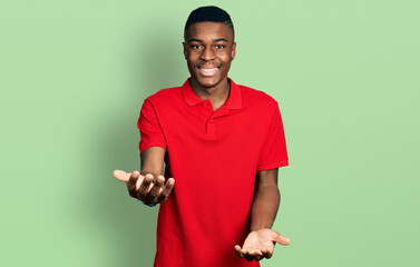 Young african american man wearing casual red t shirt smiling cheerful with open arms as friendly welcome, positive and confident greetings