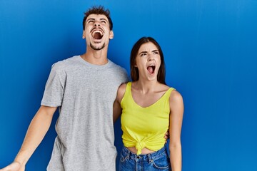 Young hispanic couple standing together over blue background angry and mad screaming frustrated and furious, shouting with anger. rage and aggressive concept.