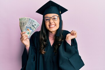 Young hispanic woman wearing graduation uniform holding usa dollars screaming proud, celebrating victory and success very excited with raised arm