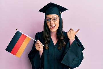 Young hispanic woman wearing graduation uniform holding germany flag pointing thumb up to the side smiling happy with open mouth