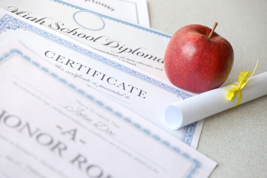 A honor roll recognition, certificate of achievement and high school diploma lies on table with small scroll and red apple. Education documents