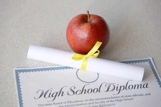 A high school diploma lies on table with small scroll and red apple. Education documents