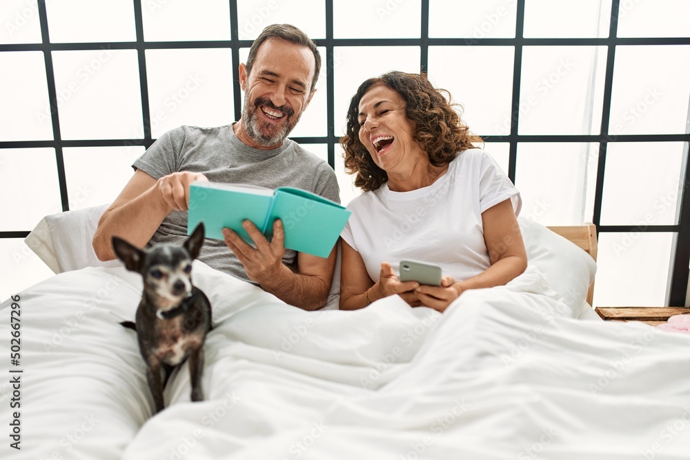 Sticker middle age hispanic couple reading book and using smartphone. lying on the bed with dog at home.