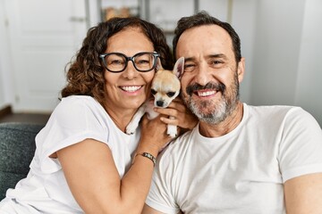 Middle age hispanic couple smiling happy sitting on the sofa holding dog at home.