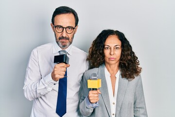 Middle age couple of hispanic woman and man holding reporter microphones thinking attitude and...