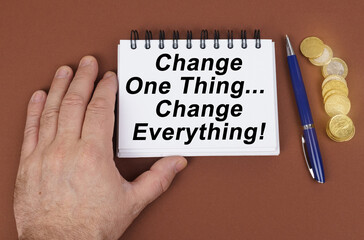 On a brown surface are coins, a pen and a notepad with the inscription - Change One Thing and...