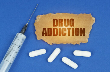 On a blue surface lie a syringe, pills and a cardboard sign with the inscription - Drug Addiction