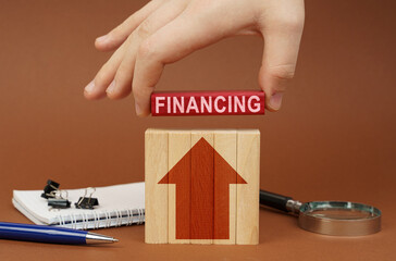 On a brown surface are office items, wooden blocks, in the hand is a red block with the inscription - Financing