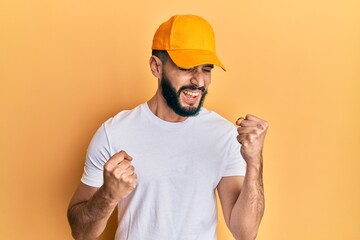 Young man with beard wearing yellow cap celebrating surprised and amazed for success with arms...