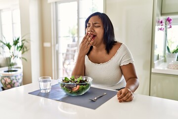 Obraz na płótnie Canvas Young hispanic woman eating healthy salad at home bored yawning tired covering mouth with hand. restless and sleepiness.