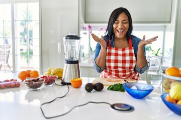 Young hispanic woman making healthy smoothie celebrating mad and crazy for success with arms raised...