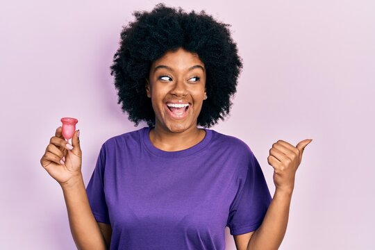 Young african american woman holding menstrual cup pointing thumb up to the side smiling happy with open mouth