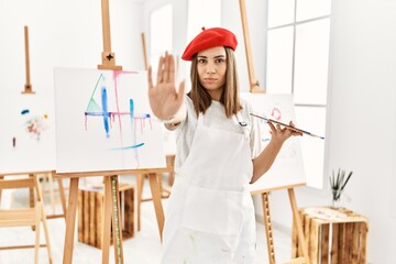 Young artist woman painting on a canvas at art studio doing stop sing with palm of the hand. warning expression with negative and serious gesture on the face.