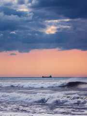 Seascape with a small container ship in the horizon crossing the Atlantic Ocean in Portugal and a dramatic sunset sky with dark blue clouds. View from the coast. Vertical composition