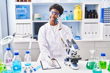 African american woman wearing scientist uniform talking on the smartphone write on clipboard at laboratory