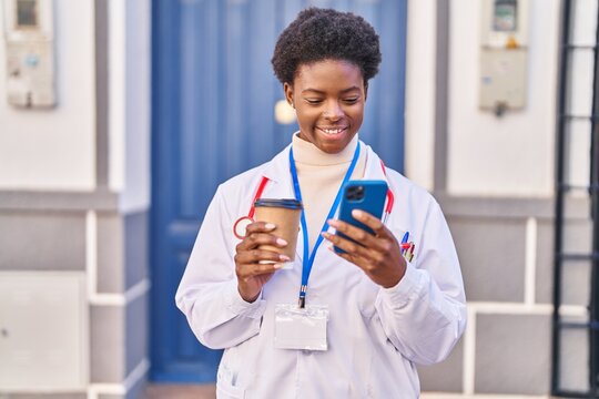 African american woman wearing doctor uniform using smartphone drinking coffee at street