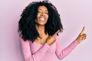 African american woman with afro hair wearing casual pink shirt smiling and looking at the camera pointing with two hands and fingers to the side.