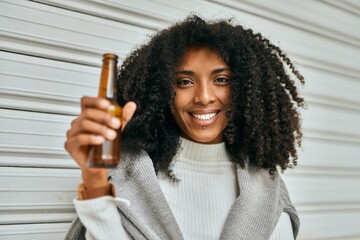 Young african american woman smiling happy holding bottle of beer at the city.