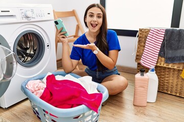 Young brunette woman doing laundry using smartphone pointing aside with hands open palms showing...