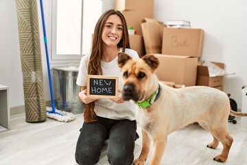 Young hispanic woman holding new home blackboard sitting on floor with dog at new home