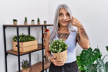 Middle age grey-haired woman holding green plant pot at home pointing with hand finger to face and nose, smiling cheerful. beauty concept