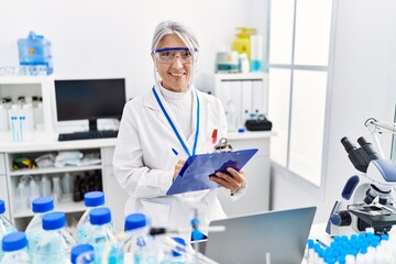 Middle age grey-haired woman wearing scientist uniform writing on clipboard working at laboratory